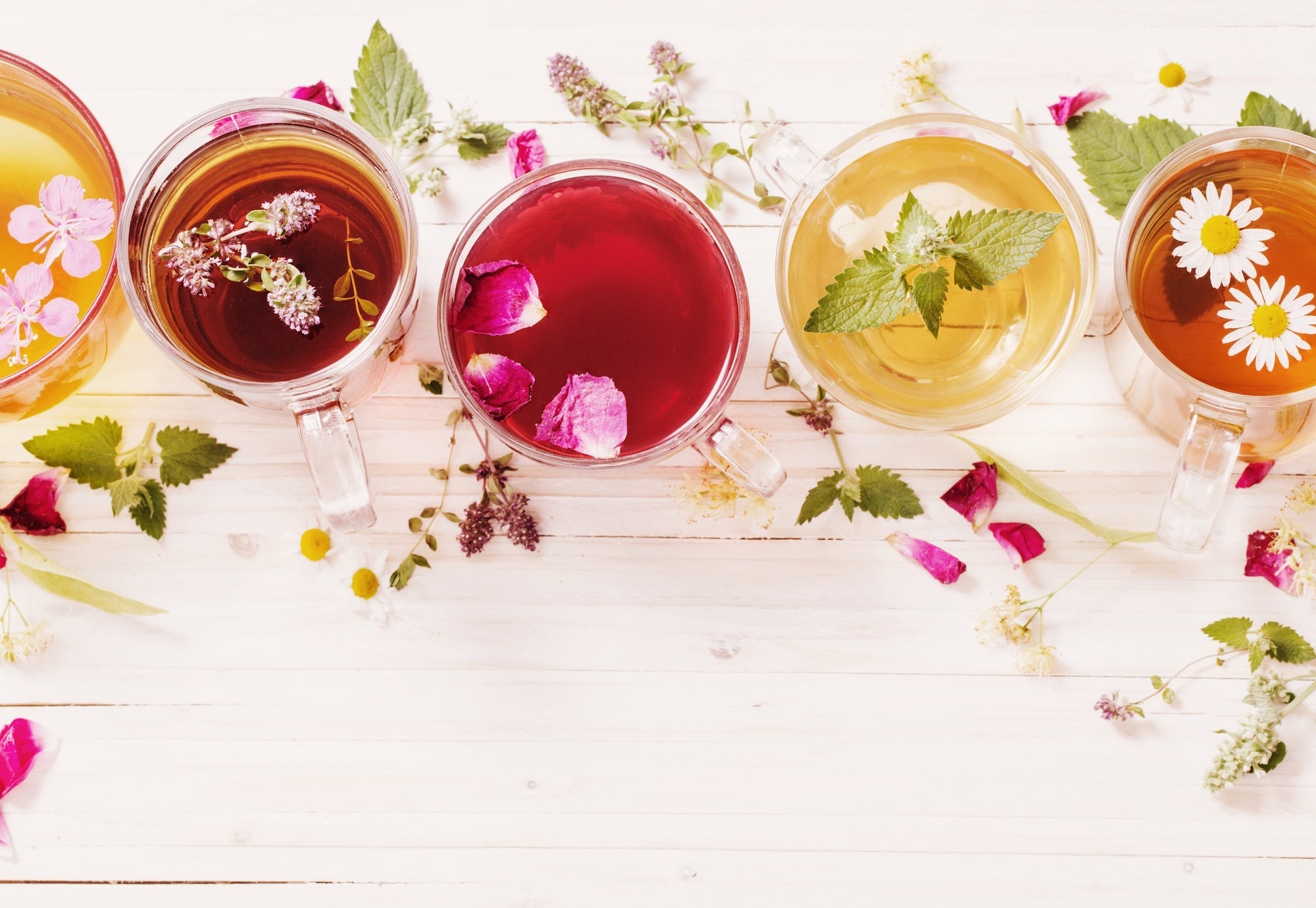 A variety of colorful herbal teas in glass mugs, each infused with different flowers and herbs, displayed on a white wooden surface with scattered fresh leaves and petals.