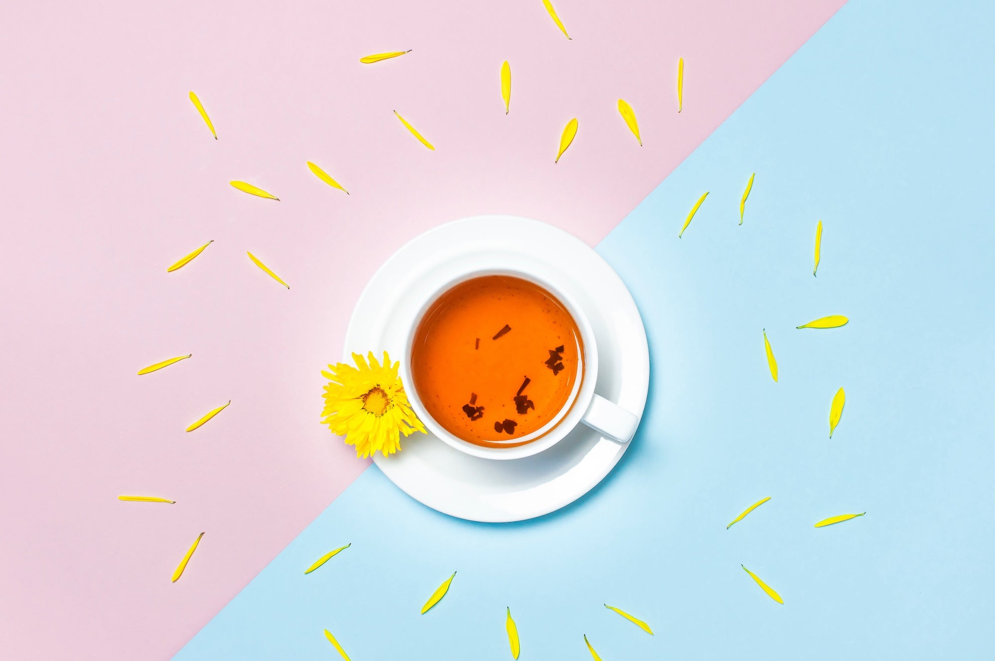 A white teacup filled with golden herbal tea, adorned with a bright yellow chrysanthemum flower, sits on a pastel pink and blue split background.