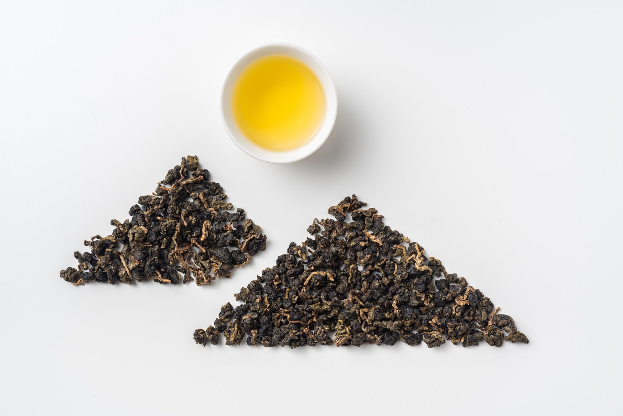 A cup of golden oolong tea alongside two piles of loose-leaf tea arranged neatly on a white background, showcasing its quality and texture.