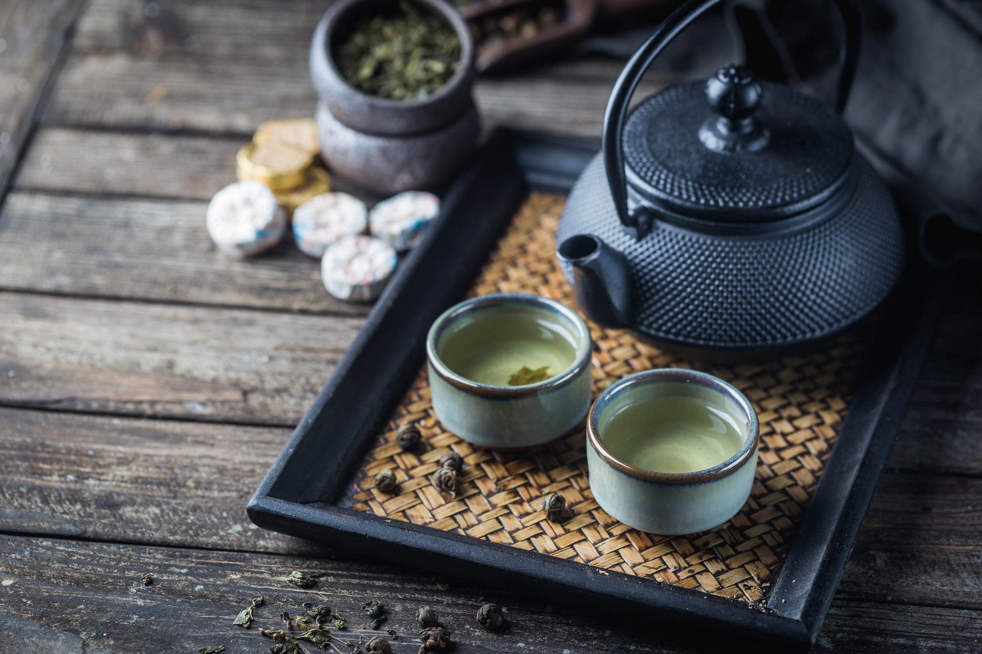 A traditional black cast iron teapot with two rustic ceramic cups filled with Chinese green tea, placed on a woven bamboo tray. Loose tea leaves and compressed tea cakes are scattered on a rustic wooden table in the background.