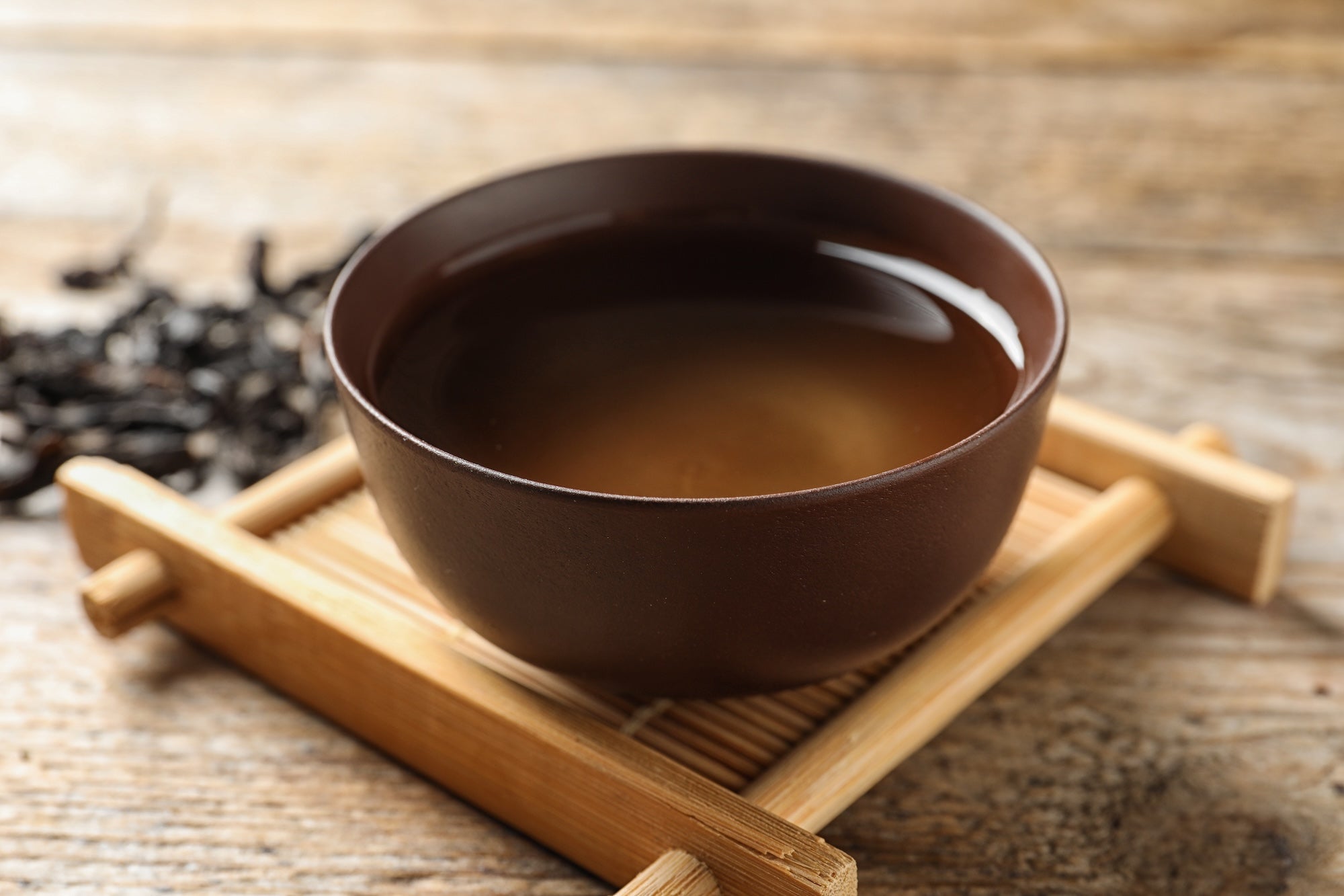 A traditional ceramic bowl filled with freshly brewed Wuyi oolong tea, placed on a bamboo tray. The tea's rich amber color reflects its deep roasting process, with loose Wuyi rock tea leaves scattered in the background.