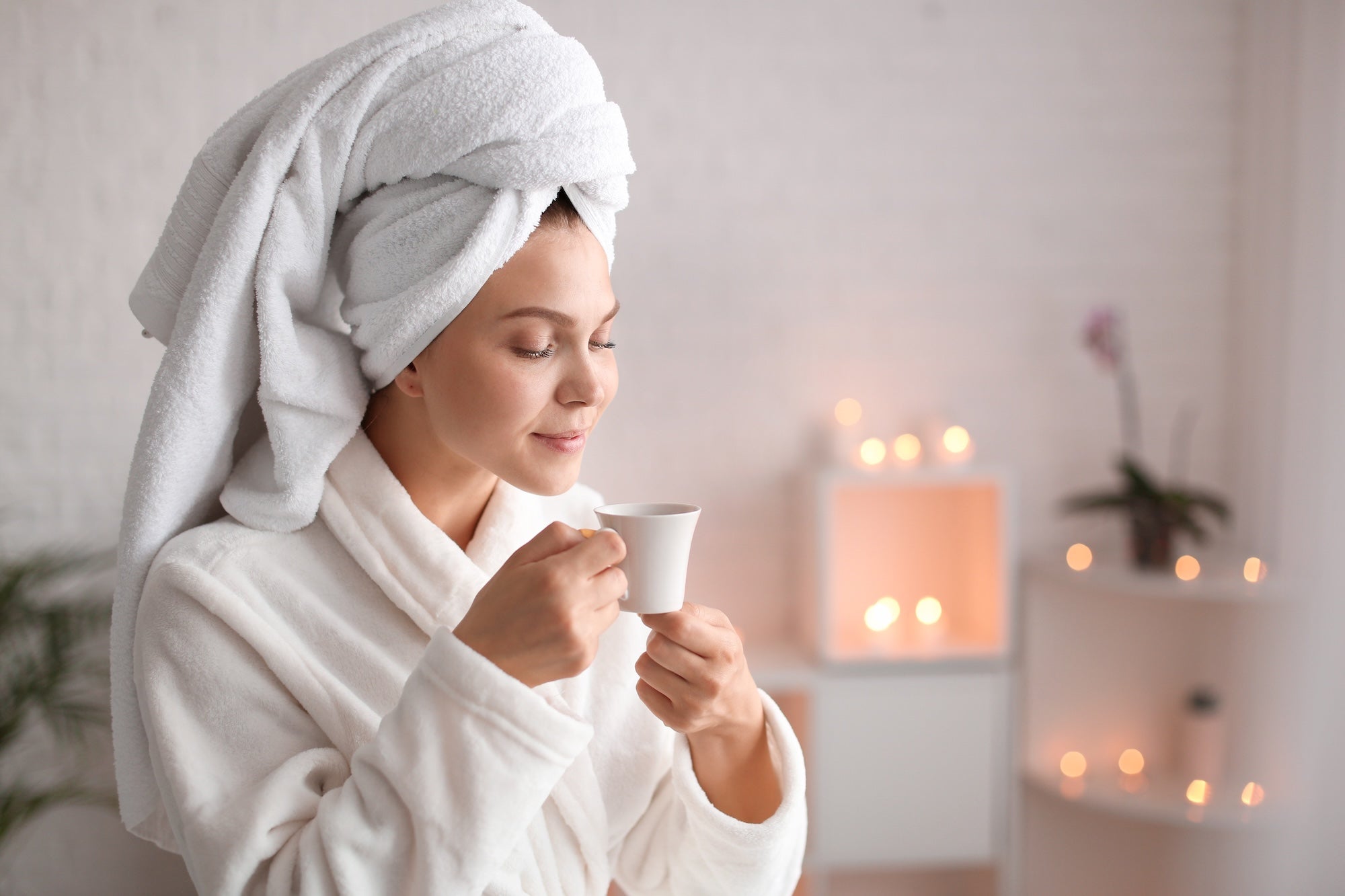 A woman in a white bathrobe with a towel wrapped around her head, enjoying a warm cup of tea in a spa-like setting with soft candlelight, symbolizing relaxation and the skin-enhancing benefits of tea.