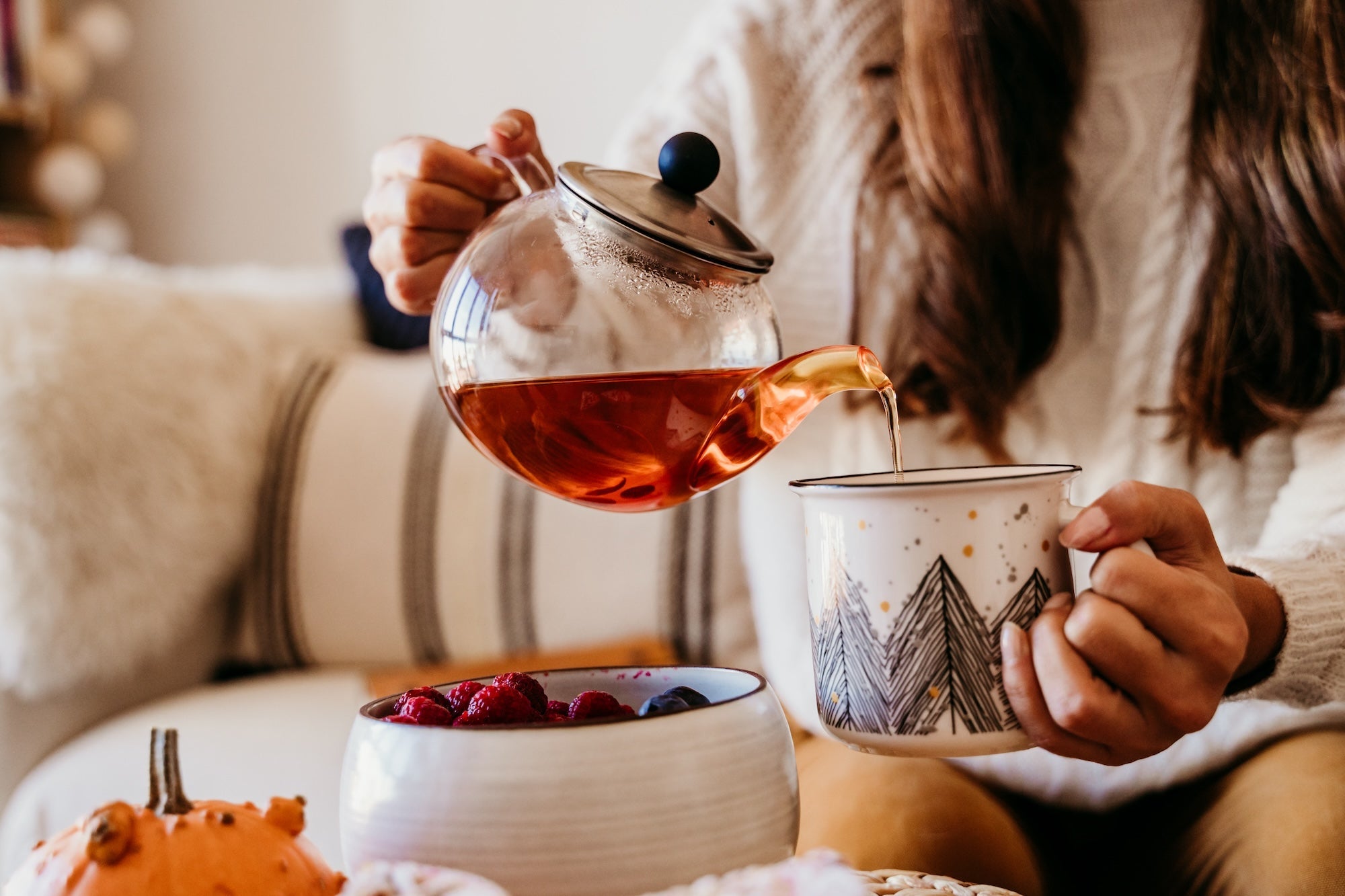 A person pouring tea from a glass teapot into a cozy mug.