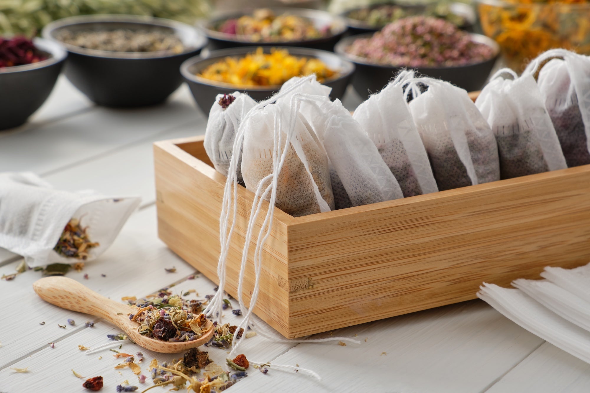 A wooden box filled with neatly arranged organic tea bags, surrounded by dried herbal tea ingredients in bowls and a wooden spoon with loose tea leaves on a white wooden surface.