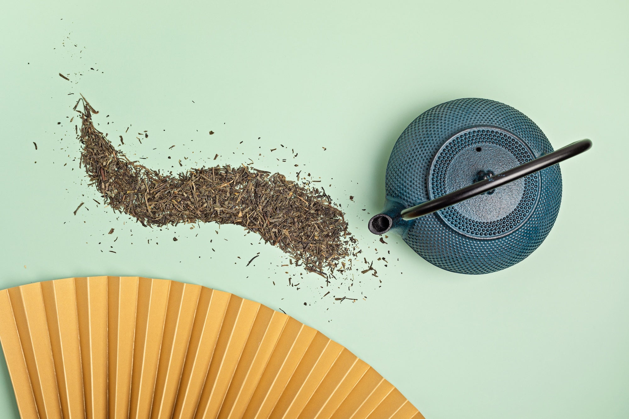 A traditional cast iron teapot with a curved handle, spilling loose-leaf tea onto a pastel green background. The tea leaves form an artistic, flowing pattern. A gold-colored folding fan is partially visible in the foreground, adding an elegant touch.