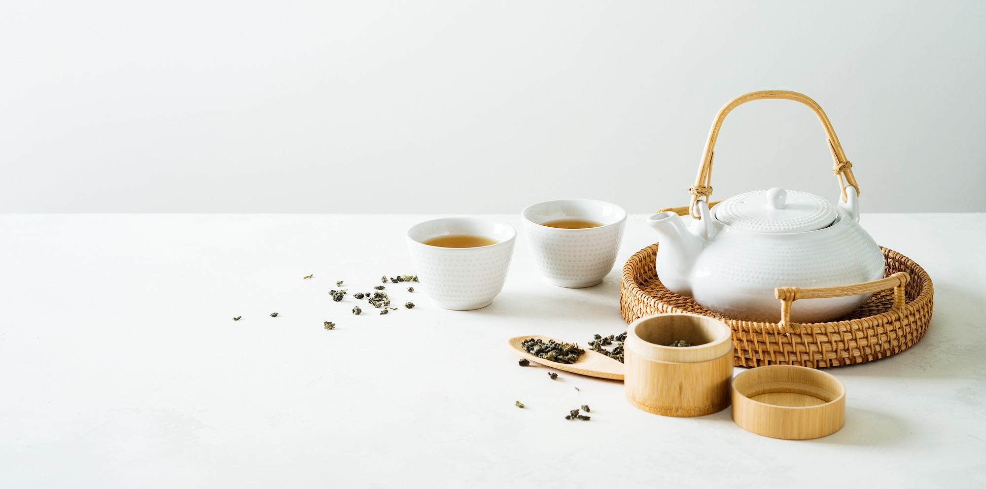 A minimalist tea setup featuring a white ceramic teapot with a woven handle, two teacups filled with golden tea, and a bamboo tea scoop with loose tea leaves, set against a clean white background.