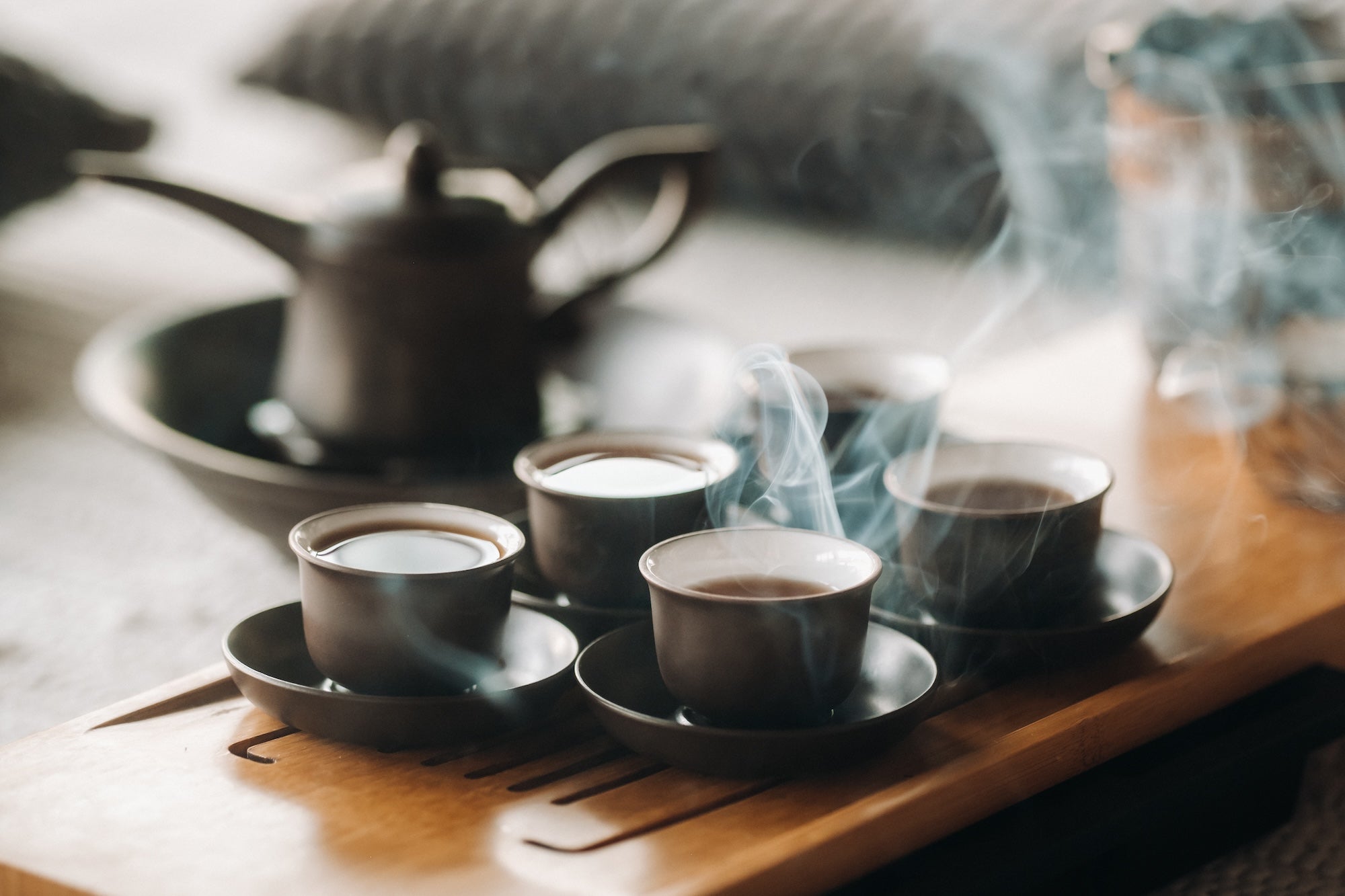 A set of black ceramic tea cups filled with steaming hot tea sits on a wooden tea tray, with a traditional teapot in the blurred background. The warm, elegant setting evokes a peaceful tea ceremony atmosphere.