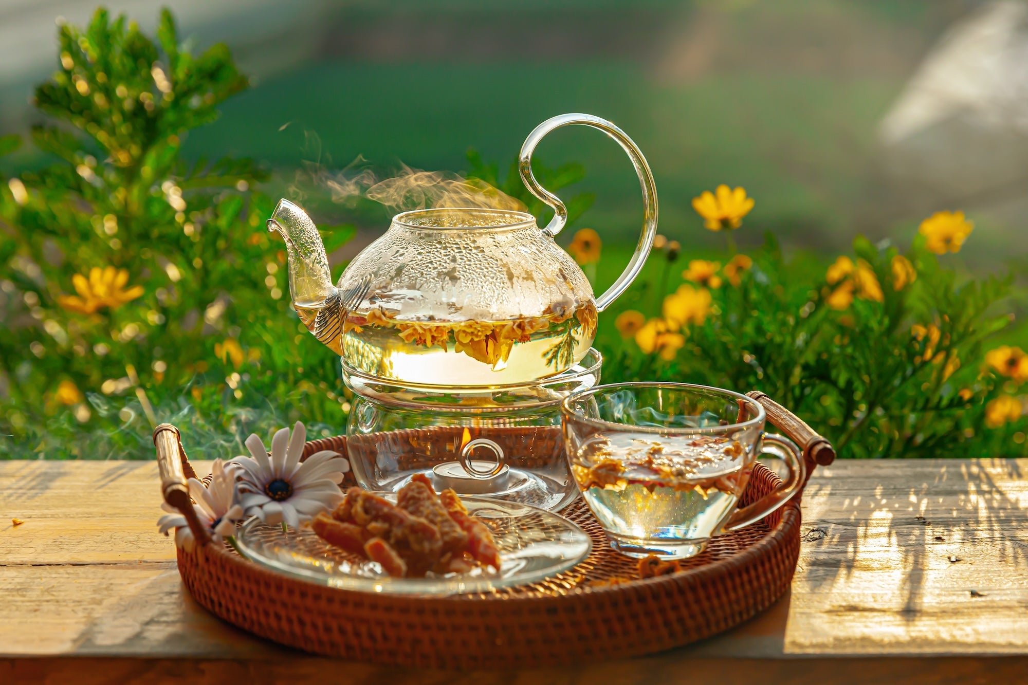 A glass teapot filled with hot chrysanthemum tea sits on a woven tray alongside a glass teacup, dried flowers, and snacks. The setting is an outdoor wooden table with blooming yellow flowers and green foliage in the background, bathed in warm sunlight.