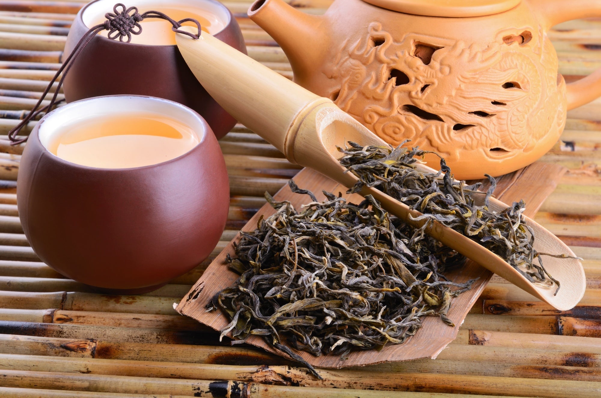 A traditional Chinese tea setup featuring a clay teapot with intricate dragon carvings, two rustic tea cups filled with golden-hued tea, and a bamboo scoop with loose dried tea leaves, all arranged on a bamboo mat.