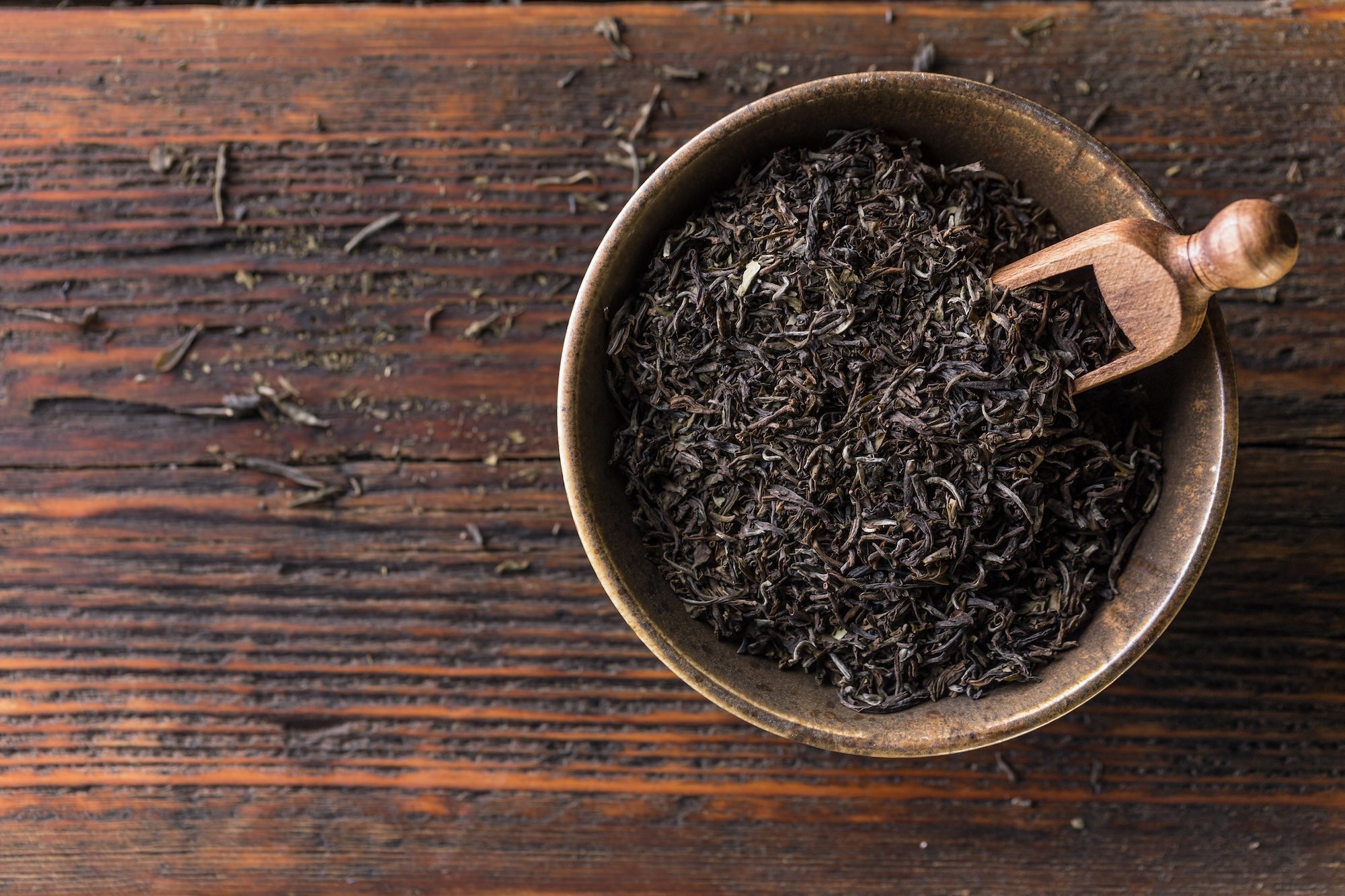 A rustic bowl filled with loose-leaf black tea, accompanied by a small wooden scoop, placed on a weathered wooden surface. The dark, finely twisted tea leaves highlight the rich aroma and natural caffeine content of premium black tea.