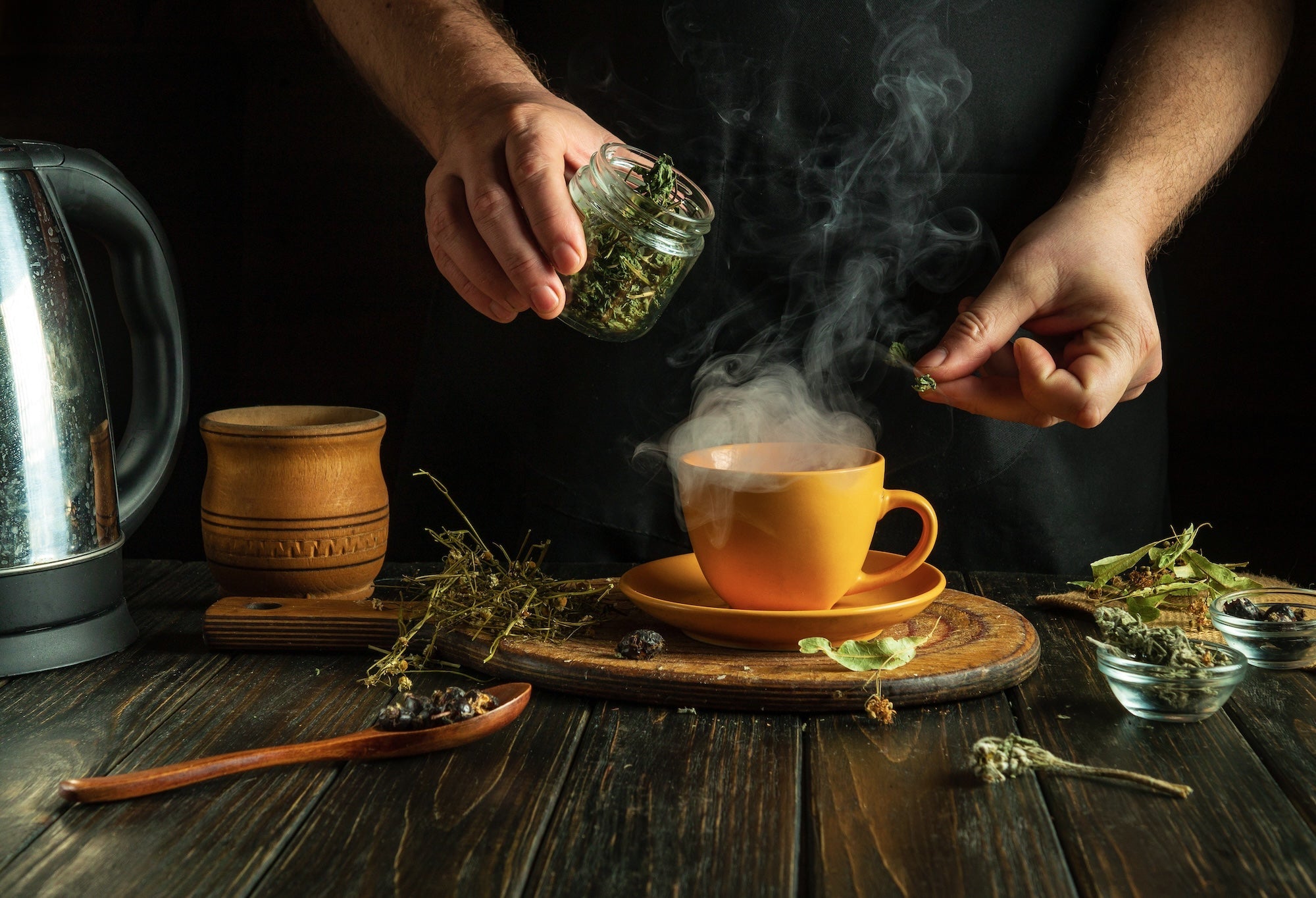 A person in a dark apron prepares herbal tea, adding loose dried herbs from a glass jar into a steaming yellow cup. The rustic wooden table holds a kettle, a wooden mug, dried herbs, and a wooden spoon with berries, creating a warm and cozy tea-making atm